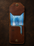 A brown leather card holder wallet with a top flap and card slots, shown open with cash and cards inside on a dark textured background. The wallet has brass snap buttons and an embossed 'EX' on the front.