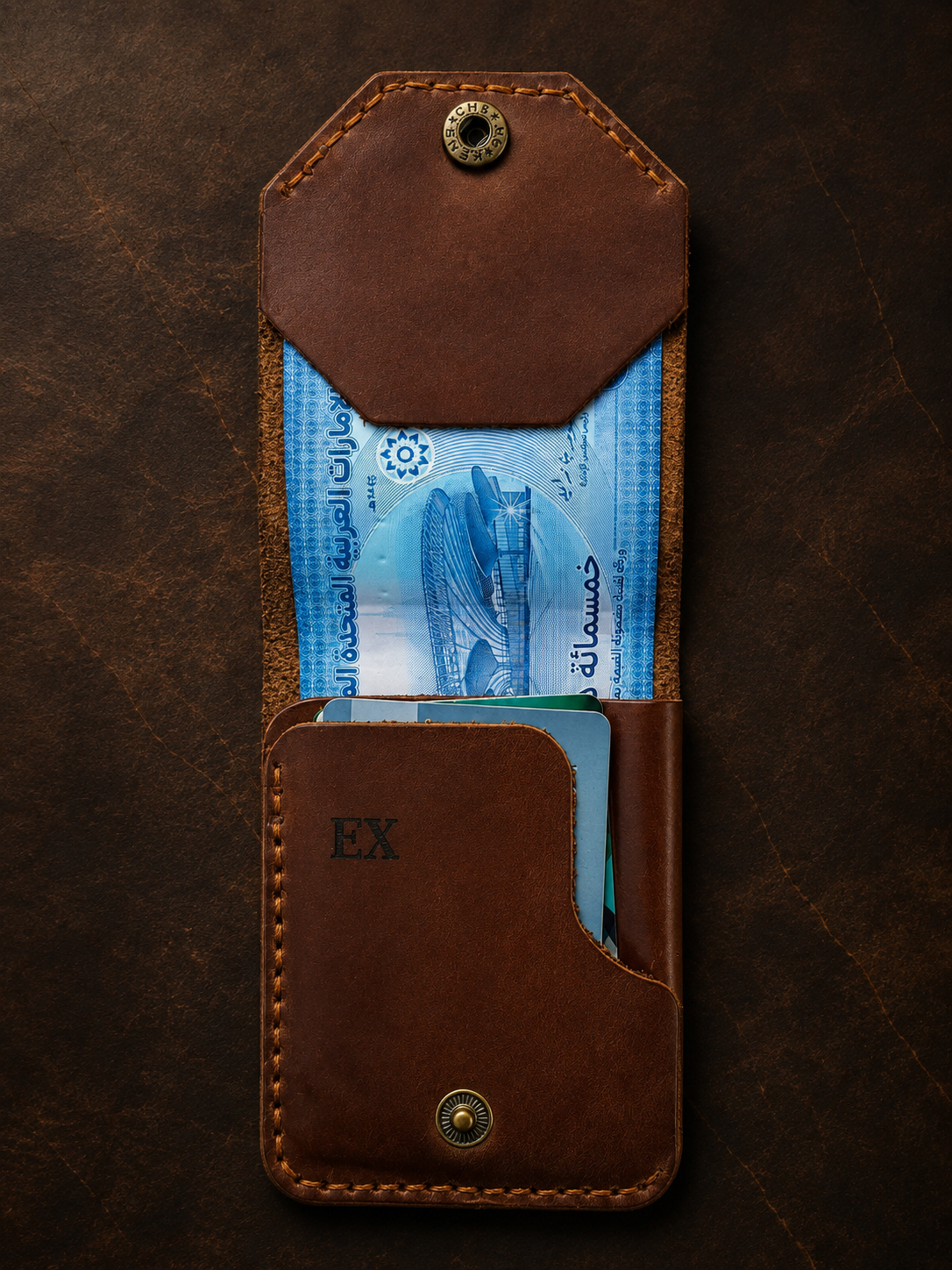 A brown leather card holder wallet with a top flap and card slots, shown open with cash and cards inside on a dark textured background. The wallet has brass snap buttons and an embossed 'EX' on the front.