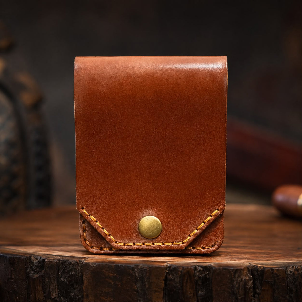 A brown leather card holder or small wallet with a snap button closure, displayed on a rustic wooden surface against a dark blurred background.